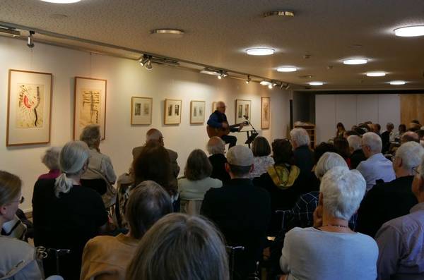 Uli Führe spielt Gitarre vor großem Publikum im Foyer der Katholischen Akademie. Im Hintergrund hängen Kunstwerke von Bruno Schley.