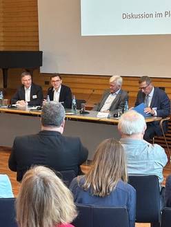 Quelle: Katholische Akademie der Erzdiözese Freiburg Vier Personen sitzen am Tisch in der Aula, im Vordergrund sind einige sitzende Teilnehmende von hinten zu sehen.