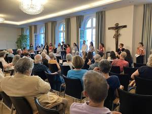 Quelle: Katholische Akademie der Erzdiözese Freiburg Etwa 50 Personen sitzen in deinem Saal mit einem großen Kreuz an der Wand und hören zu, wie die Gesprächspartner vorgestellt werden, diese stehen vorne in einer Reihe.