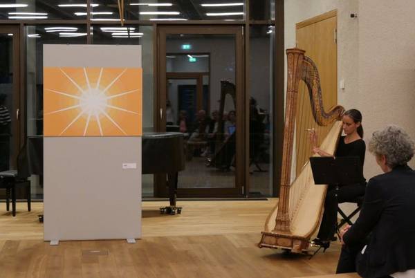 Quelle: Katholische Akademie der Erzdiözese Freiburg Links im Bild ein orange-gelbes Bild mit einem strahlenden sonnenähnlichen Kreis in der Mitte, daneben sitzt eine junge Frau und spielt die Harfe.